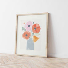 Load image into Gallery viewer, an art print of an illustration of colourful poppies in a vase framed in a pale wooden frame leaning against a plain white wall