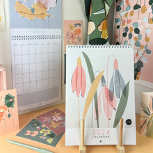 Load image into Gallery viewer, The front of a floral 2024 wall calendar propped up in a stand in the foreground. An open calendar hanging in the background with various floral and botanical postcards and cards pinned up in the background or scattered on the table top. There is a botanical tea towel and poster hanging in the background  

