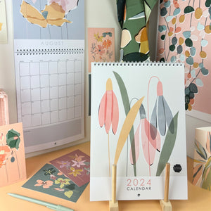 The front of a floral 2024 wall calendar propped up in a stand in the foreground. An open calendar hanging in the background with various floral and botanical postcards and cards pinned up in the background or scattered on the table top. There is a botanical tea towel and poster hanging in the background  