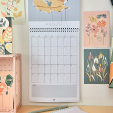 Load image into Gallery viewer, A floral 2024 wall calendar hanging opened on the month of august. Various botanical postcards are pinned up next to it.
