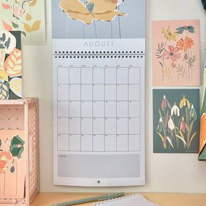 A floral 2024 wall calendar hanging opened on the month of august. Various botanical postcards are pinned up next to it.