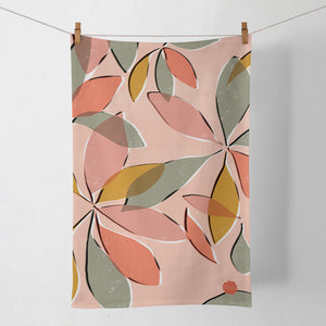 A coral coloured tea towel hanging by pegs on a washing line against a pale background. The tea towel features an abstract botanical design of the leaves of a schefflera nora house plant.