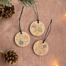 Load image into Gallery viewer, Decorative circular tags with floral patterns hanging on a textured surface with pine cones and string lights.
